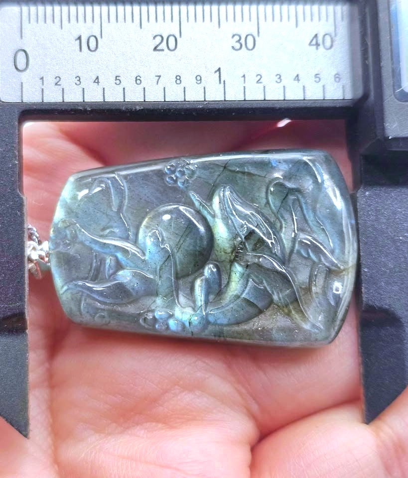 A hand holds a polished labradorite pendant with a carved dragon design, positioned under a caliper to show its size. The pendant exhibits characteristic blue-green iridescent flashes from the labradorite stone.
