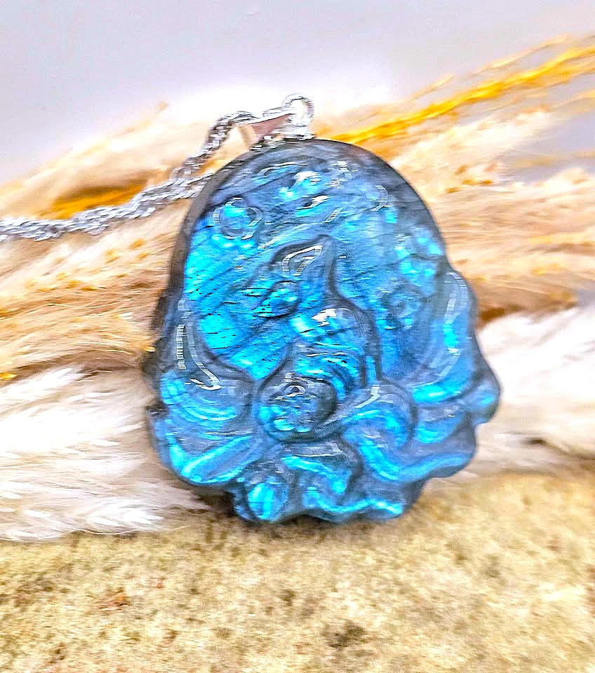 A silver pendant necklace featuring a highly polished, iridescent blue stone with a floral relief design, resting on a textured beige surface with dried grass.
