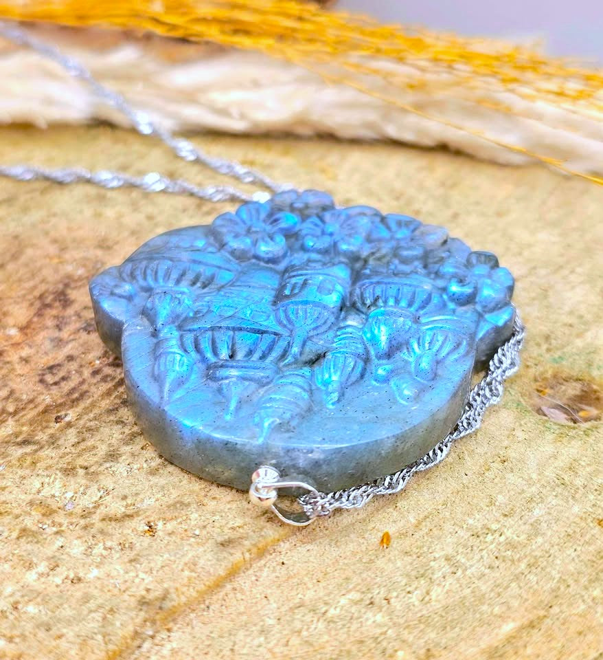 A carved blue pendant necklace with a silver chain, resting on a textured beige stone surface with dried yellow grass in the background.