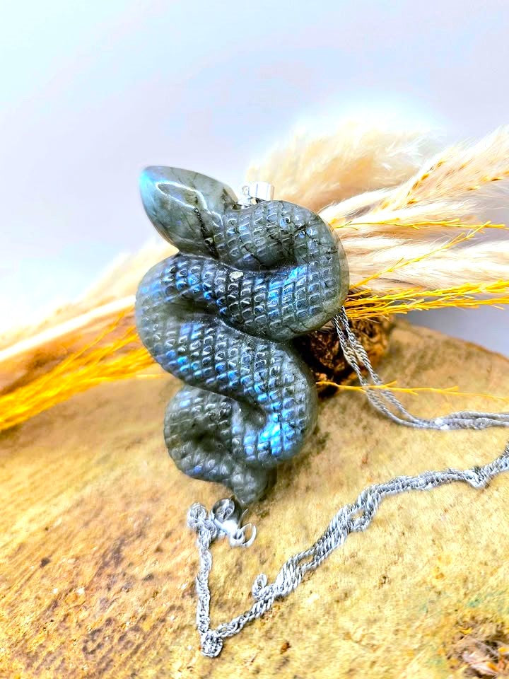 A silver snake pendant necklace with a dark blue-green iridescent labradorite finish, resting on a piece of wood with dried grass.