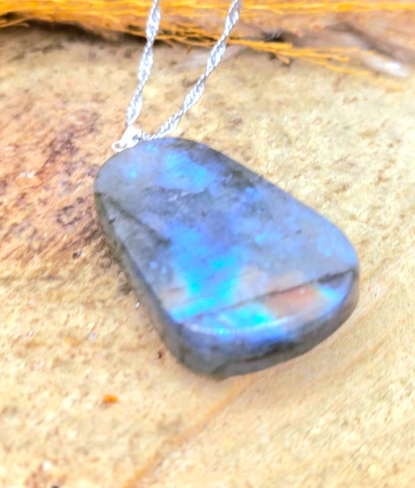 A polished labradorite stone pendant necklace with a silver chain, resting on a textured sandy surface.