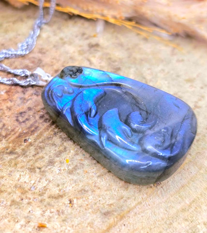 A polished blue labradorite pendant necklace with a carved wave design, resting on a textured stone surface with a silver chain.