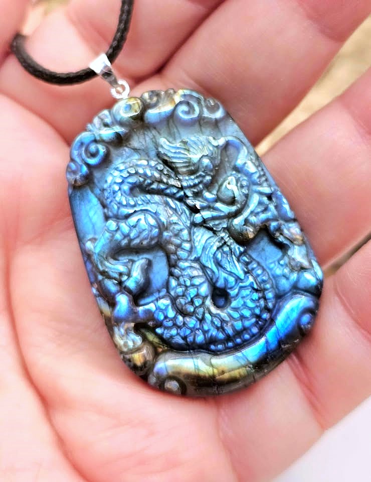 A close-up of a hand holding a carved dragon pendant necklace. The pendant is made of a blue iridescent stone with silver-toned carved details and hangs from a black cord.
