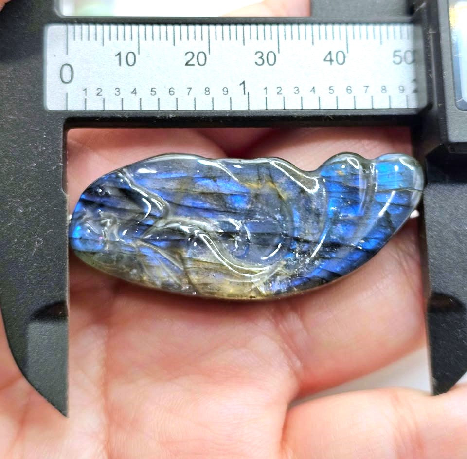 A polished labradorite stone pendant or charm resting on a person's palm, with a digital caliper above it showing its width. The stone exhibits characteristic blue and gold iridescent labradorescence.