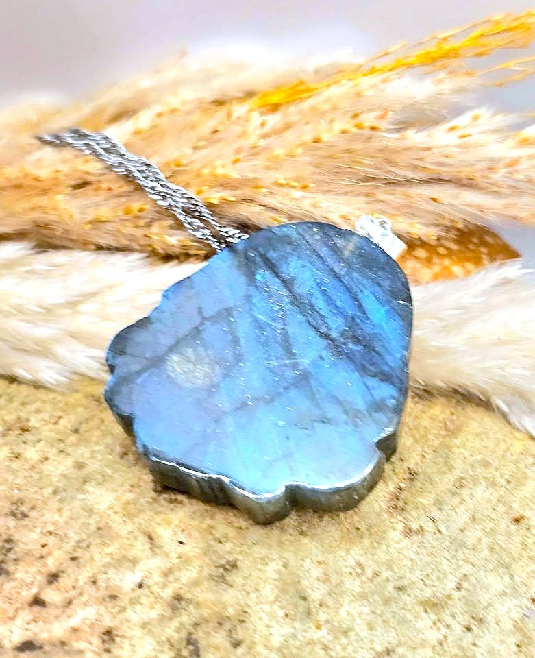 A polished labradorite stone pendant necklace with a silver chain, resting on a textured stone surface with dried grass in the background.
