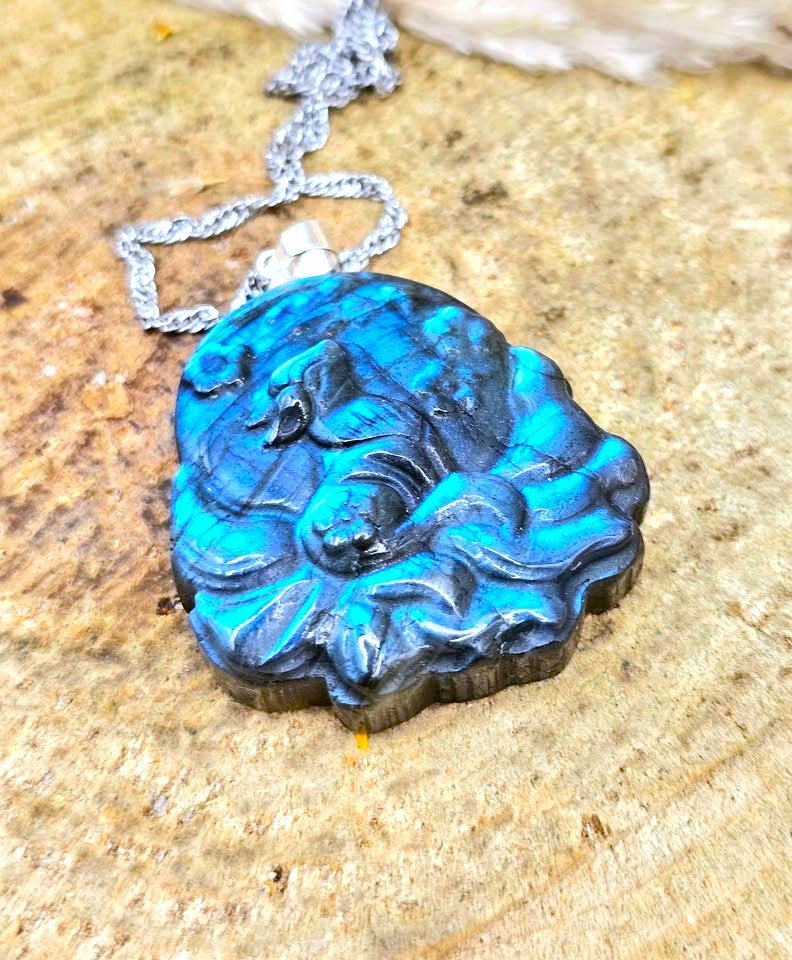 A carved labradorite pendant necklace featuring a sleeping dog design, resting on a textured stone surface. The pendant exhibits vibrant blue and black marbling, suspended from a silver-toned chain.