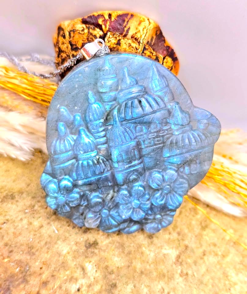A round blue carved stone pendant featuring an embossed cityscape with domes and flowers, hanging from a silver chain against a textured beige and brown background.
