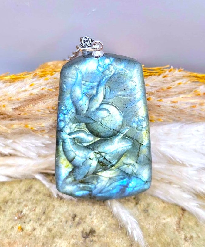 A rectangular labradorite pendant with a silver bail, featuring a carved figure holding a sphere, displayed on a textured stone surface with dried grass.
