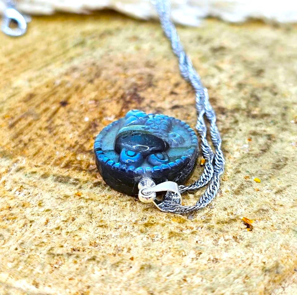A close-up of a round pendant necklace with a blue and black illustrated design, attached to a silver chain, resting on a textured beige stone surface.
