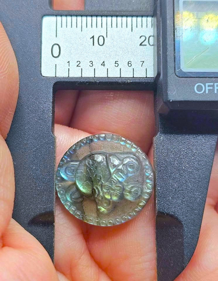 A close-up of a round, textured gray stone pendant with a floral design, held in a hand under a caliper to show its size. The pendant has a shimmering, iridescent green-blue finish.