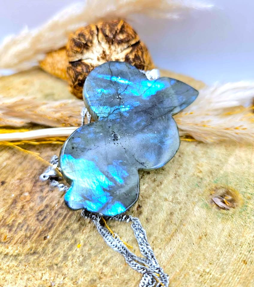 A butterfly-shaped pendant made of iridescent labradorite stone with blue and green flashes, hanging from a silver chain on a textured wood surface with dried grass.