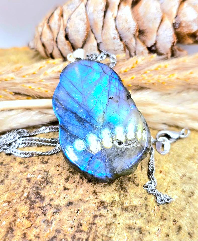 A polished labradorite pendant necklace with a blue and green iridescent surface, shown on a rustic background with dried grass and a pinecone.