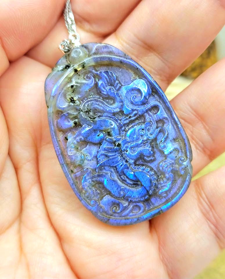 A close-up of a carved oval pendant with a blue iridescent finish and raised dragon-like motifs, held in a person's hand. The pendant has a silver bail and cord and appears to be made of a labradorite or opal-like stone.