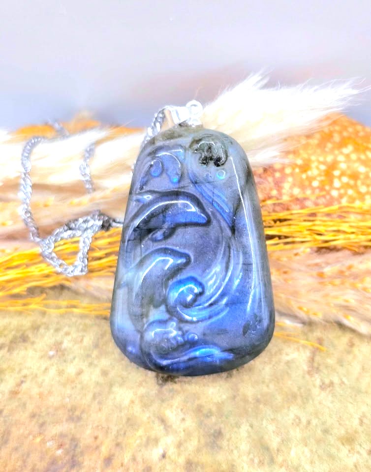A carved blue labradorite pendant necklace with a silver-toned chain, displayed on a textured surface with dried grass.
