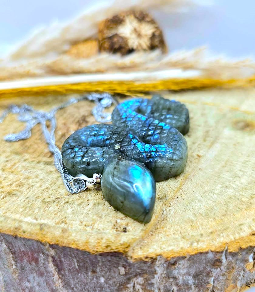 A silver-toned snake pendant necklace made from polished labradorite stones, resting on a rustic wood slice with dried grass in the background.