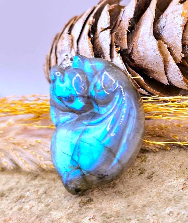 A polished labradorite heart-shaped pendant with vibrant blue and green flashes, displayed on a textured surface with a pinecone and dried grass in the background.