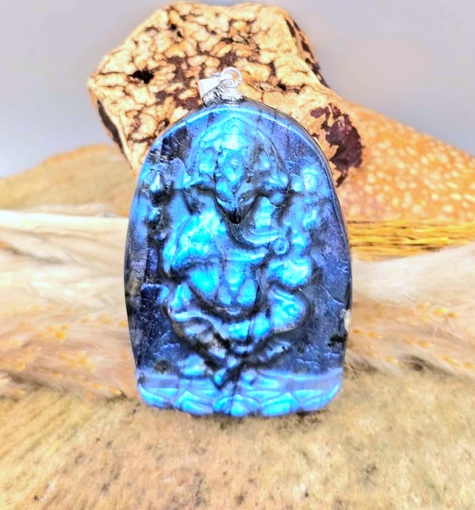 A carved blue labradorite pendant featuring a detailed Ganesha design, displayed on a piece of wood with a cork accent.