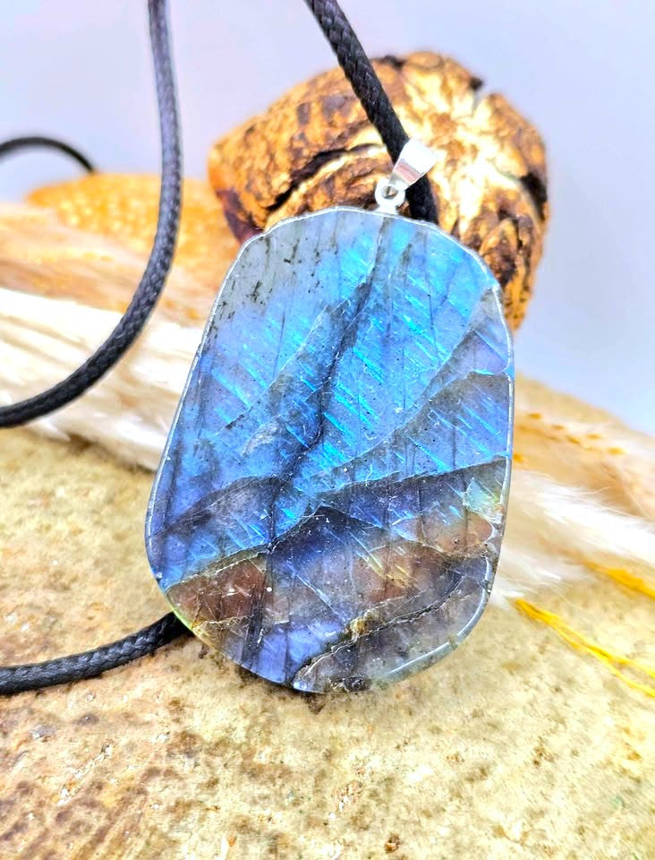 A raw labradorite stone pendant necklace with a black cord and silver bail, displayed on a textured stone surface with a piece of wood in the background.