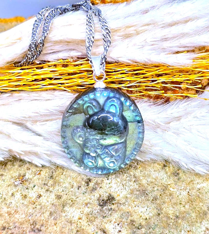 A round pendant necklace with a silver-toned chain, featuring a carved animal face design on a shimmering blue-green iridescent stone, displayed on a textured background of white fur and golden dried grass.
