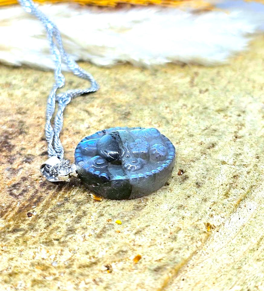 A carved oval pendant necklace made of dark blue-gray stone with a raised animal motif, hanging from a silver-toned chain on a textured sandy surface.