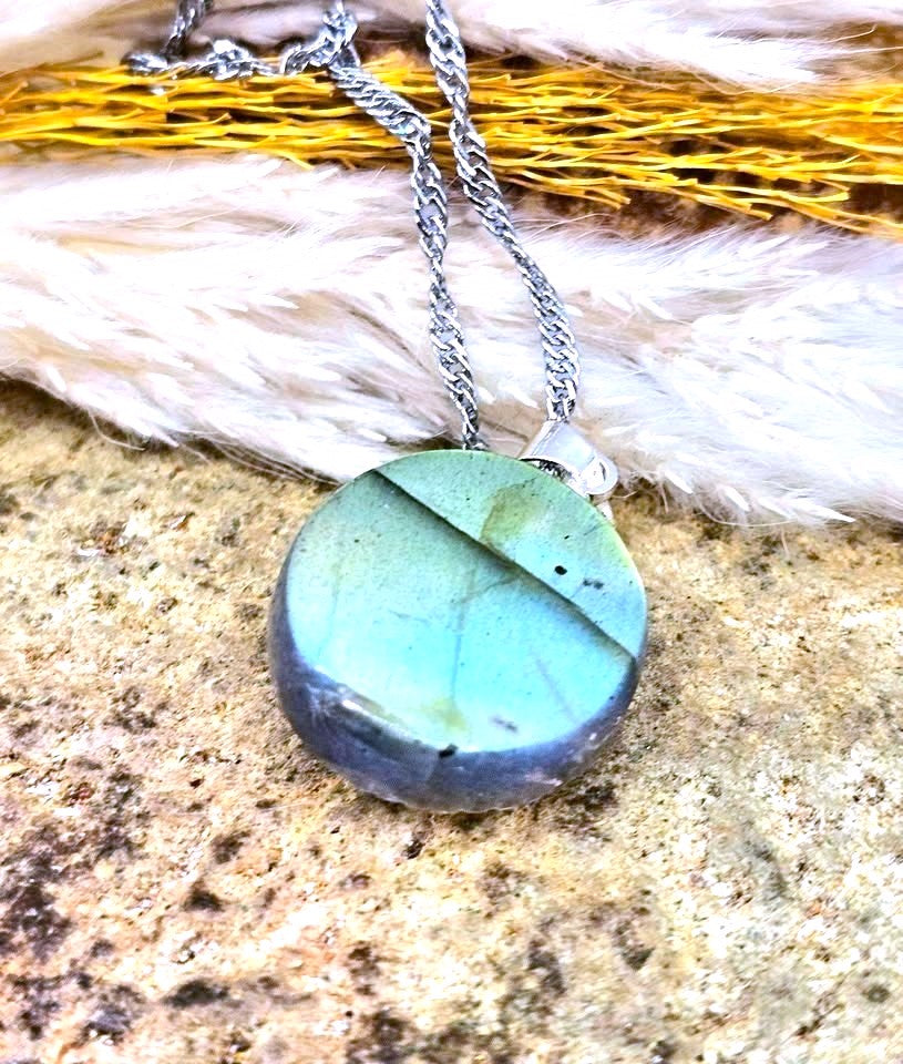 A round labradorite stone pendant necklace on a silver chain, resting on a textured stone surface with white faux fur and yellow dried grass in the background.
