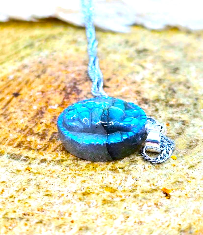 A carved oval pendant necklace made of dark blue and black stone with bright turquoise highlights, hanging on a silver-toned chain against a textured sandy background.
