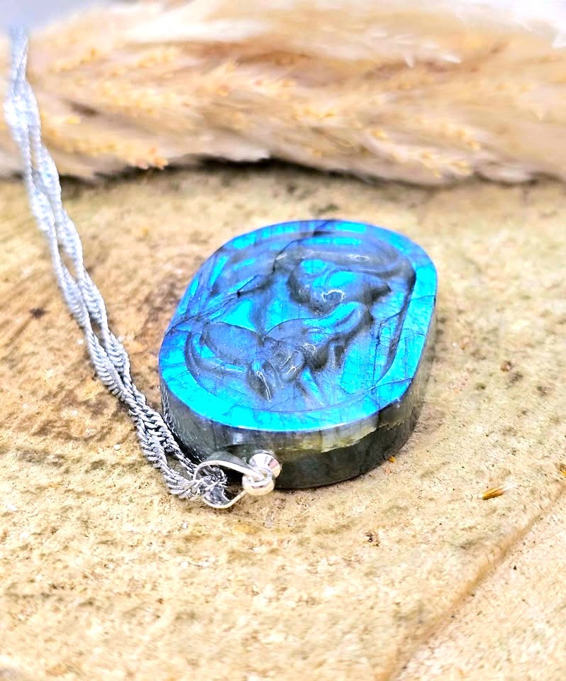 A carved oval pendant necklace made of layered turquoise and black stone, hanging from a silver chain on a textured stone surface with dried grass in the background.