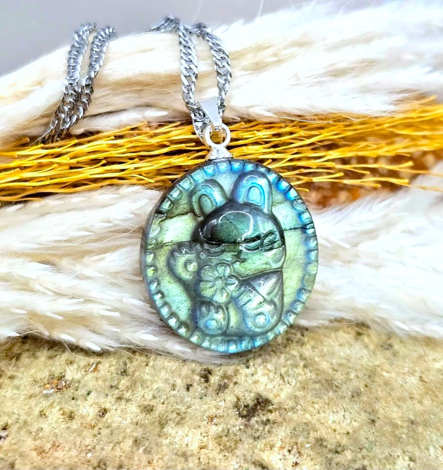 A silver chain necklace with an oval pendant featuring a stylized cartoon rabbit in green and blue iridescent stone, displayed on a textured surface with white faux fur and golden dried grass.