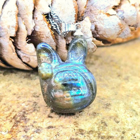 A small iridescent pendant shaped like a rabbit or bunny head, hanging from a silver loop, photographed close up against a textured stone surface with a pinecone behind it.