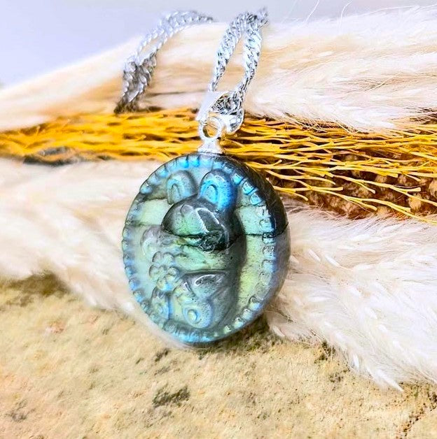 A silver chain necklace with an oval pendant featuring a carved turtle design over a blue, green, and black marbled pattern, displayed on a textured background of white fur and yellow feathers.