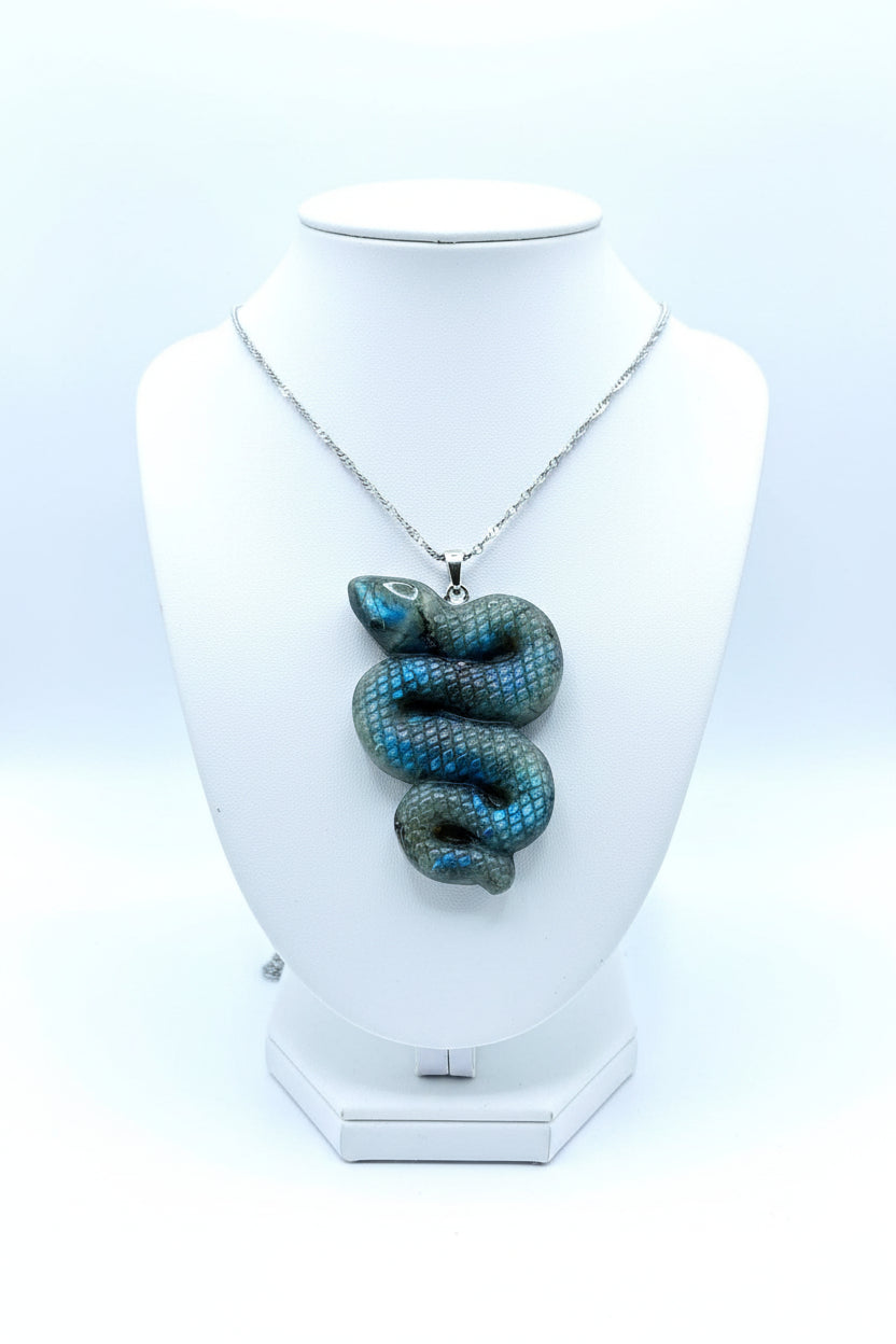 A coiled snake-shaped pendant with a textured scale pattern, featuring a blue and green iridescent finish, hanging from a silver chain on a natural wood slice background.