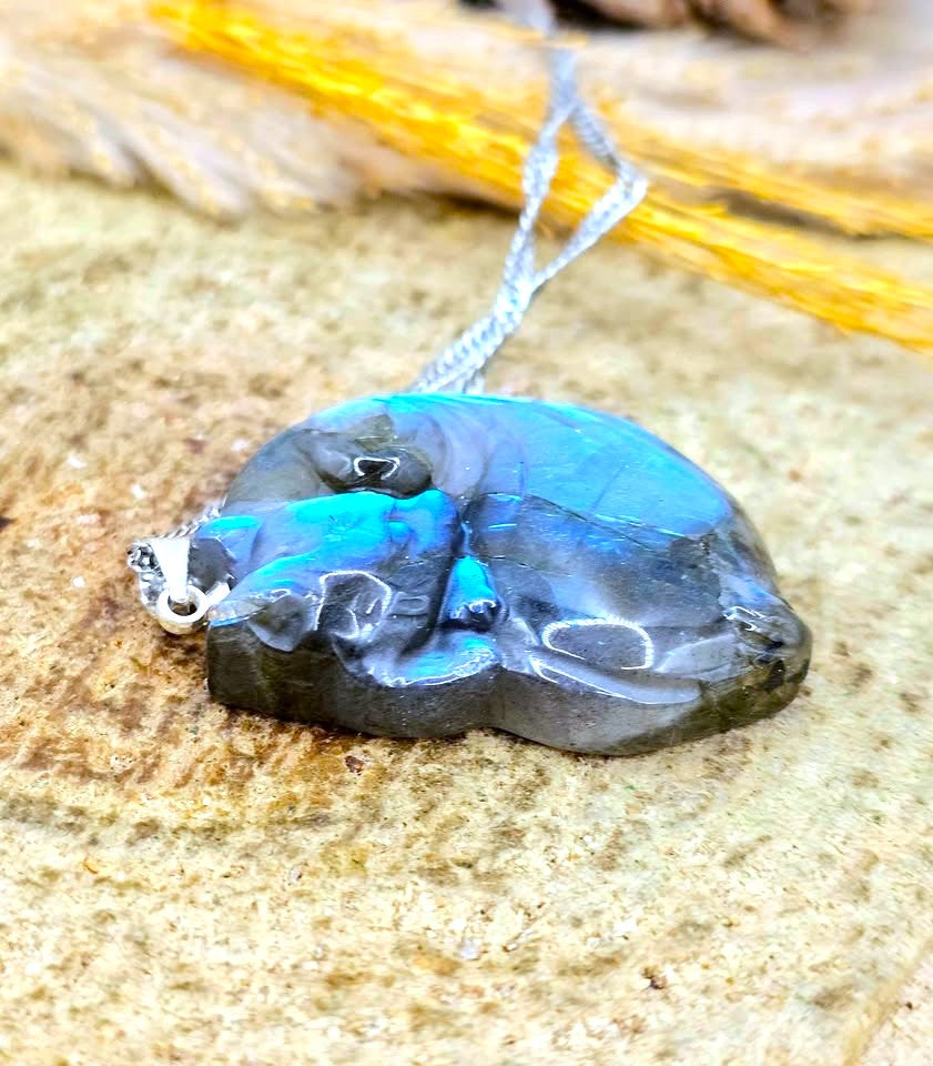 A handmade pendant made from a polished natural stone with vibrant blue and black marbled patterns, hanging from a silver chain on a textured stone surface.