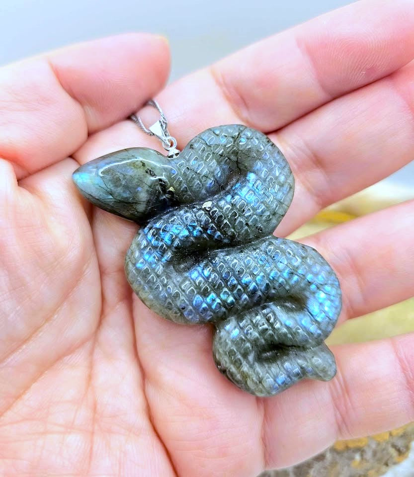 A coiled snake-shaped pendant made of iridescent labradorite stone, held in a person's hand. The pendant has a silver bail and is shown close-up against the skin of the hand.
