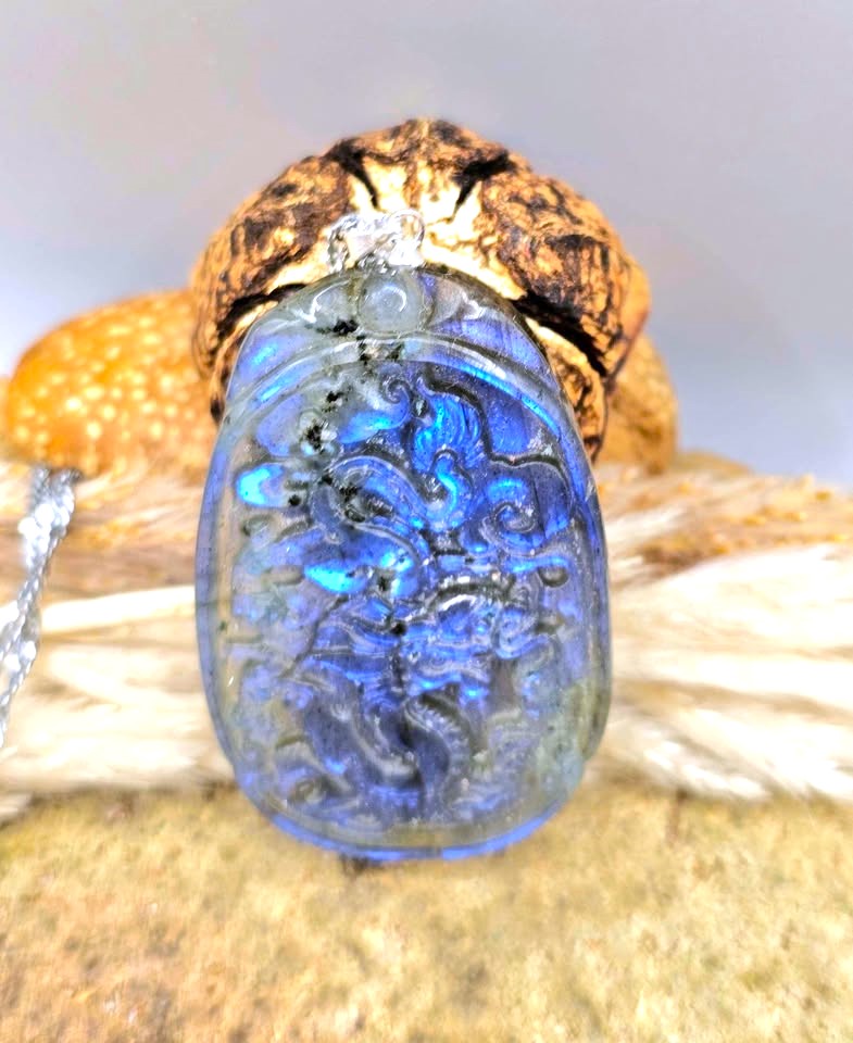 A close-up of a carved blue labradorite pendant with a gold-toned setting and a small clear stone, displayed against a neutral background.