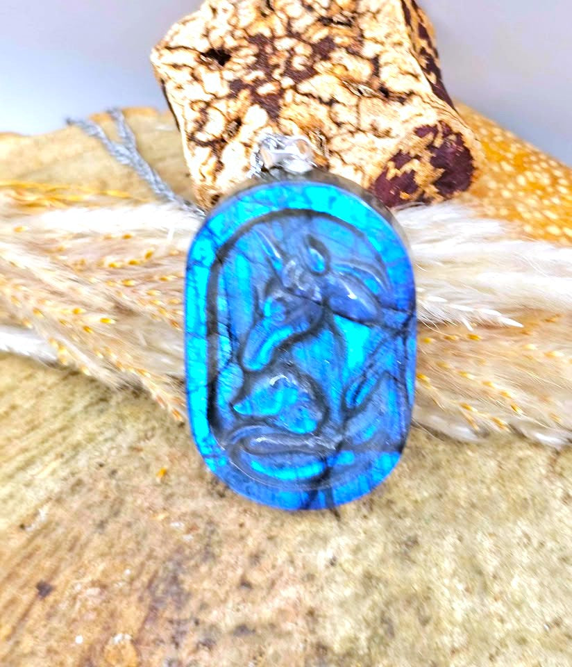 A blue agate cabochon pendant featuring an embossed unicorn design, displayed on a rustic wood and feather surface.