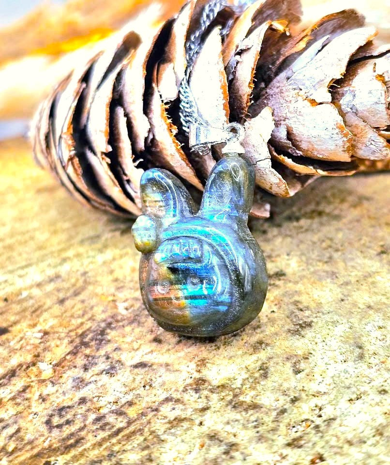 A close-up of a polished labradorite pendant shaped like a rabbit head, resting on a pinecone against a textured stone surface.