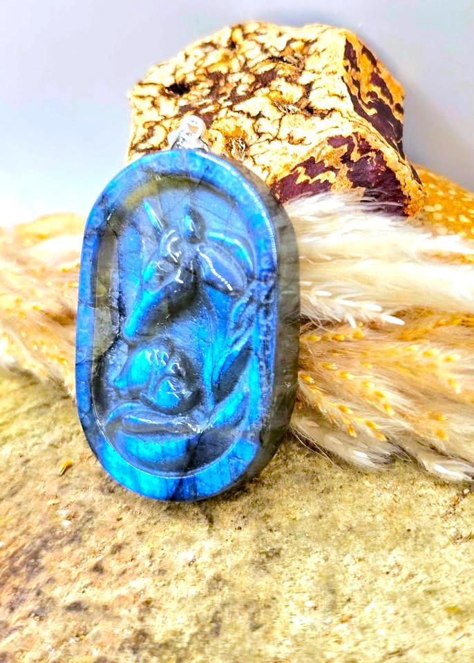 An oval cabochon pendant featuring a carved deer motif in vivid blue labradorite with dark gray and black natural banding, displayed on a textured stone surface with decorative fur and gold leaf.