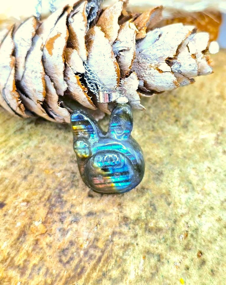 A small polished iridescent pendant or charm with a rabbit-like shape, resting on a textured stone surface beneath a pine cone.