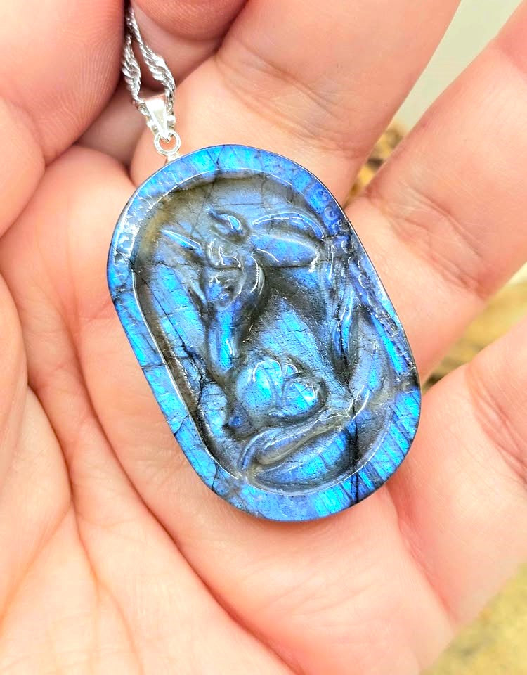 A close-up of a hand holding a carved labradorite pendant necklace. The pendant features a detailed carving of a figure, showcasing a vibrant blue iridescent labradorite edge with a dark gray polished center.