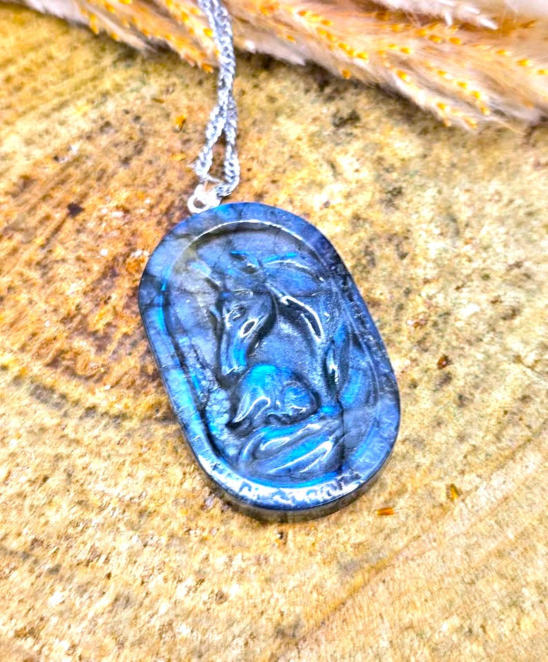 A silver necklace with an oval cabochon pendant featuring a raised horse design, set against a textured wood background with dried grass.