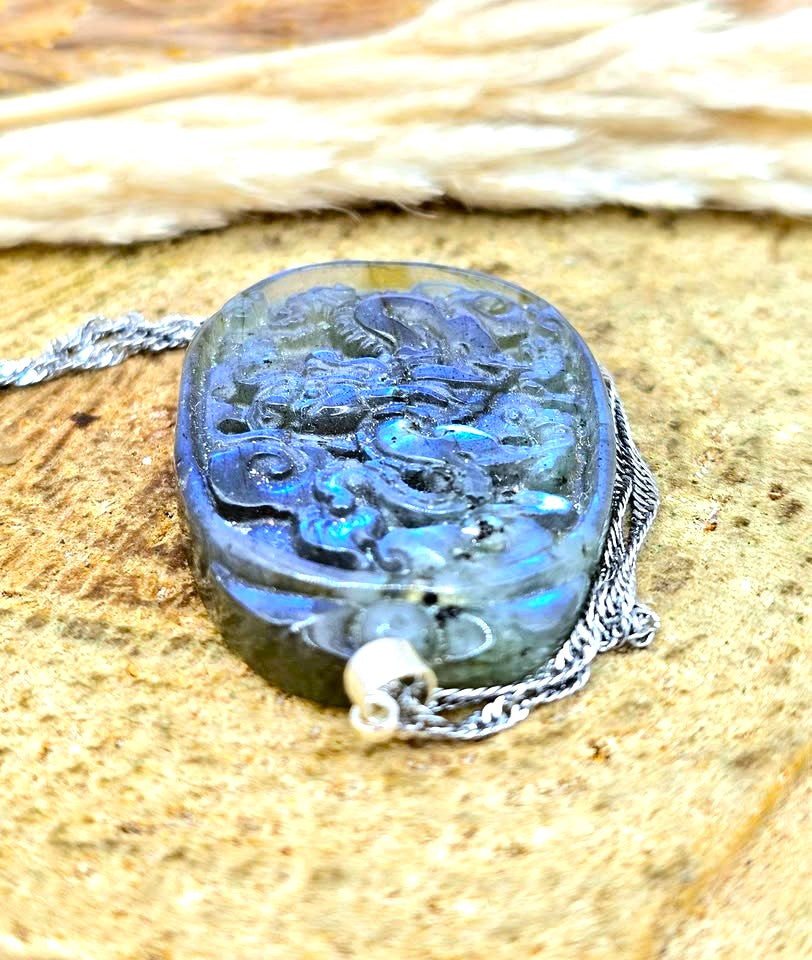 A carved oval labradorite pendant necklace with a silver chain, displayed on a textured tan surface with dried grass in the background.