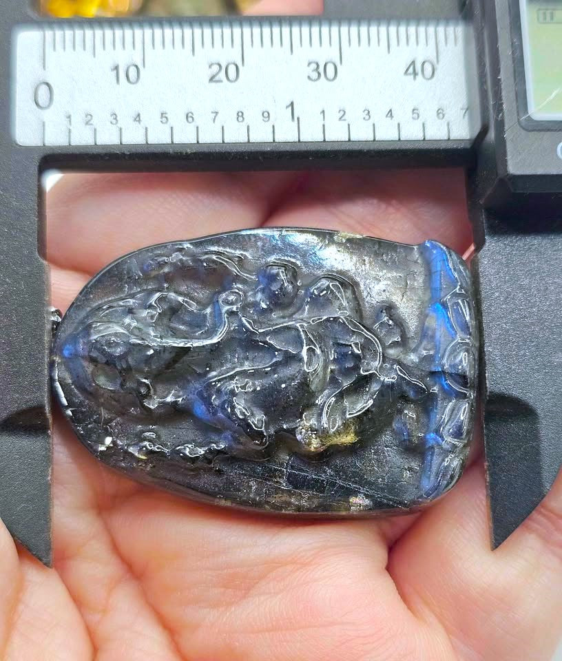A hand holds a polished dark carved stone pendant with blue flashes, positioned under a metal caliper with a ruler scale visible above it.