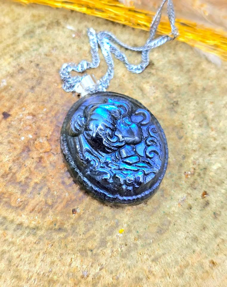 A round black carved pendant necklace with a raised face design, hanging from a silver-toned chain on a textured tan stone surface.