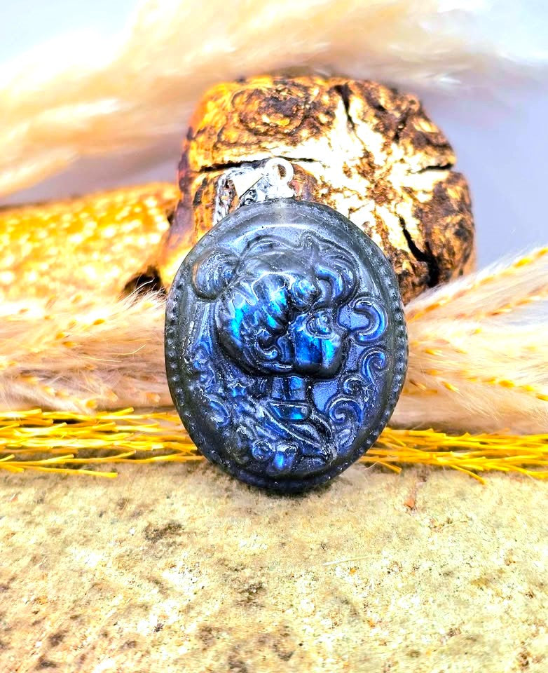 A round cabochon pendant with an embossed lion design, featuring a deep blue iridescent labradorite finish, displayed on a textured stone surface with feathers and a shell in the background.