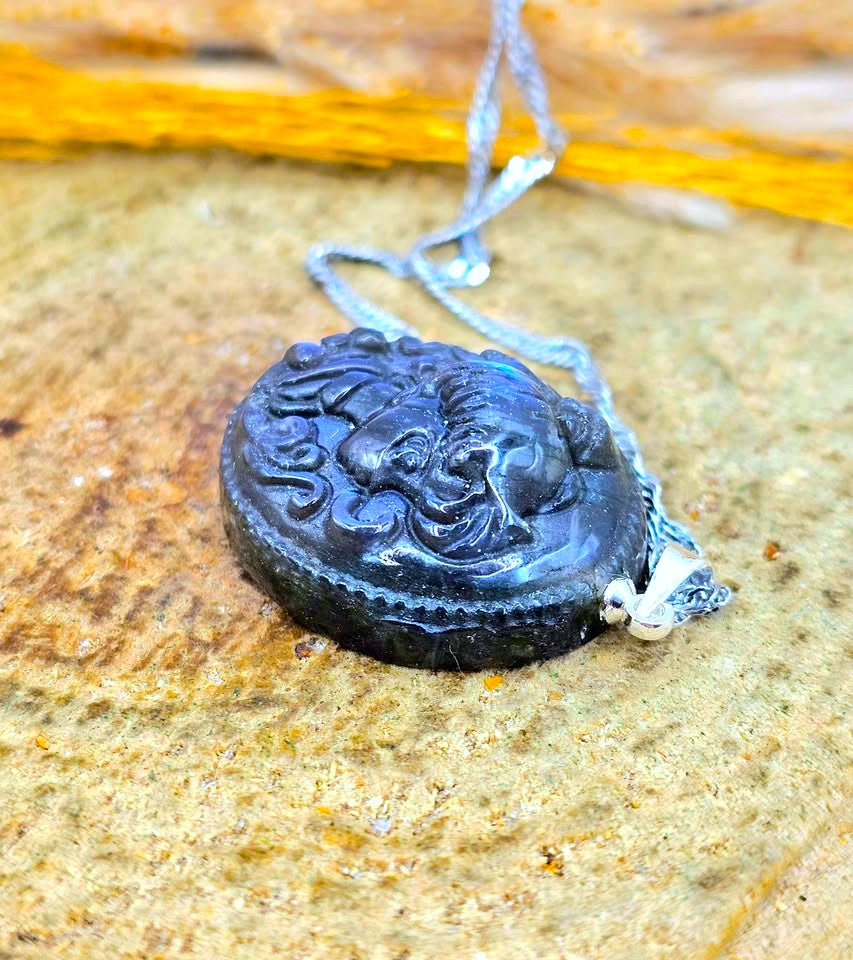 A round black carved pendant necklace with an embossed face design, hanging from a silver-toned chain on a textured sandy surface.