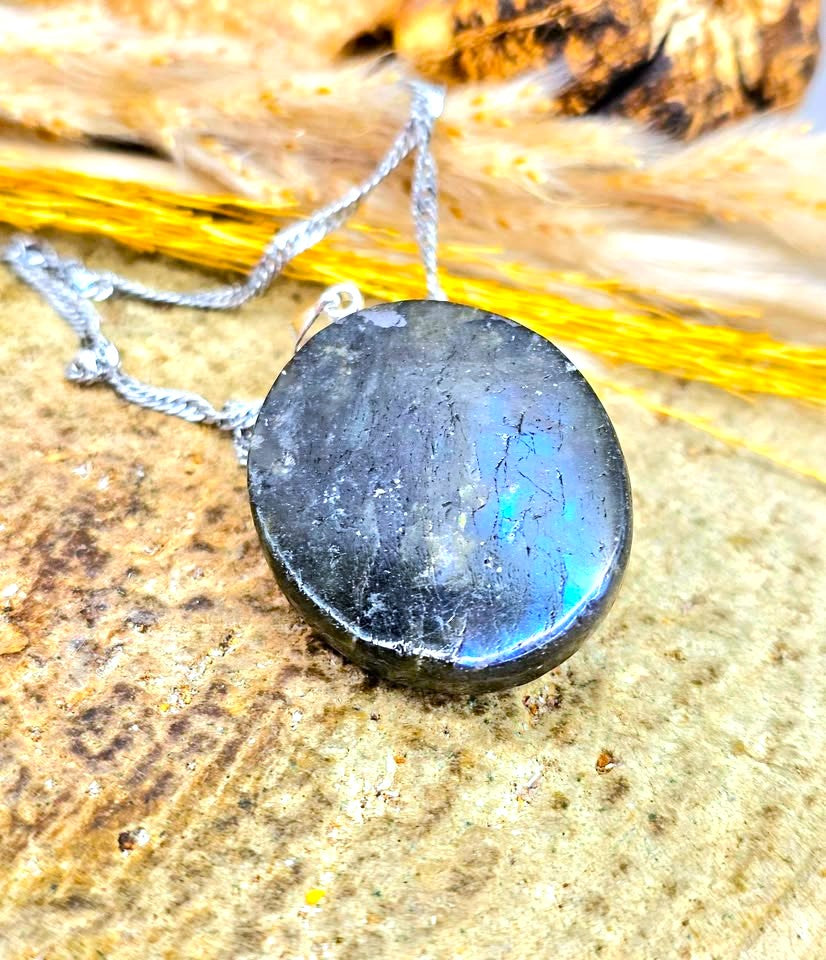 A polished round black labradorite gemstone pendant on a silver chain, displayed on a textured stone surface with dried grass in the background.