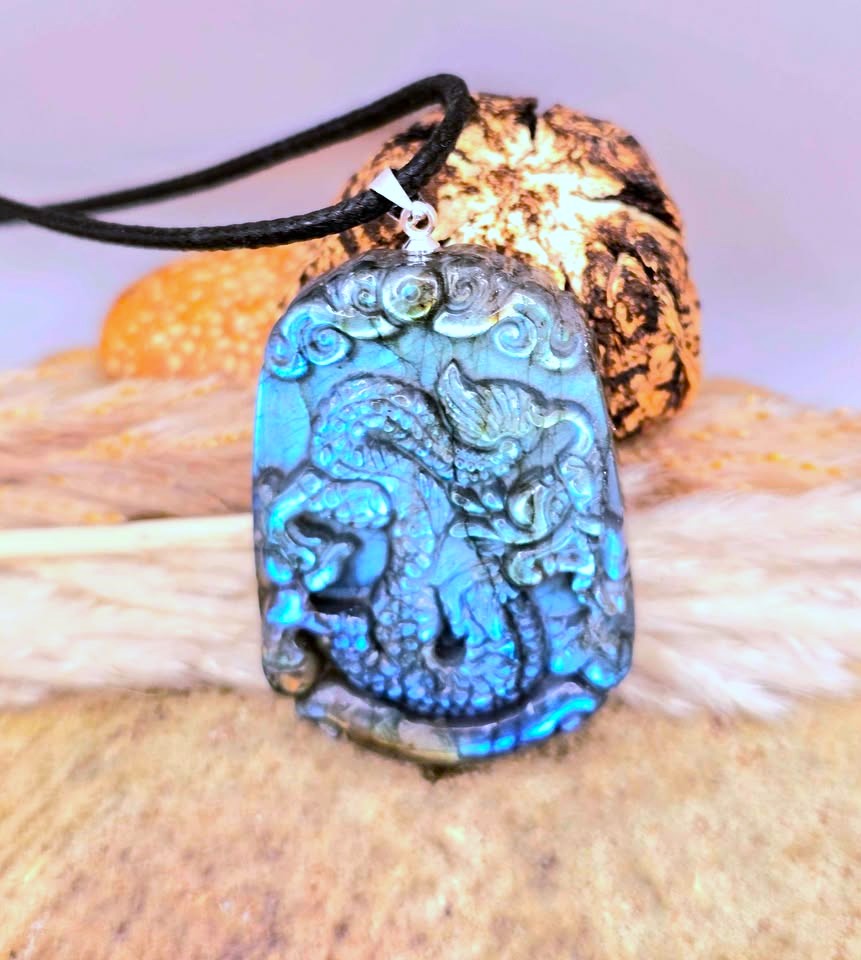 A carved dragon pendant necklace made of iridescent blue labradorite stone on a black cord, displayed against natural wood and dried grass.