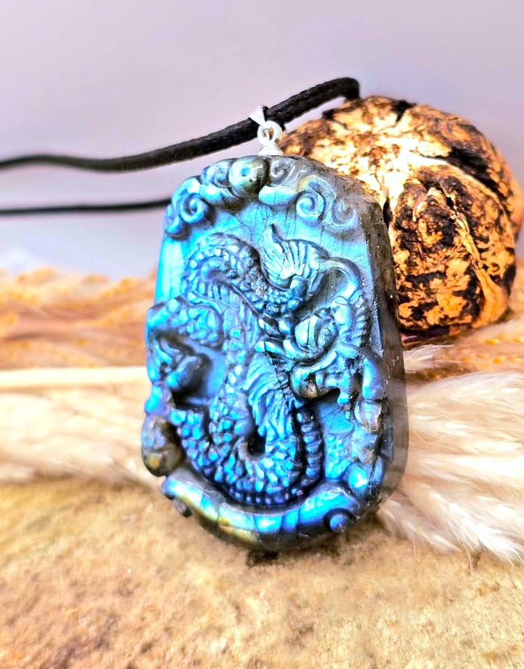 A carved dragon pendant necklace with a vibrant blue iridescent finish, hanging from a black cord. The pendant is displayed on a textured surface next to a decorative shell.
