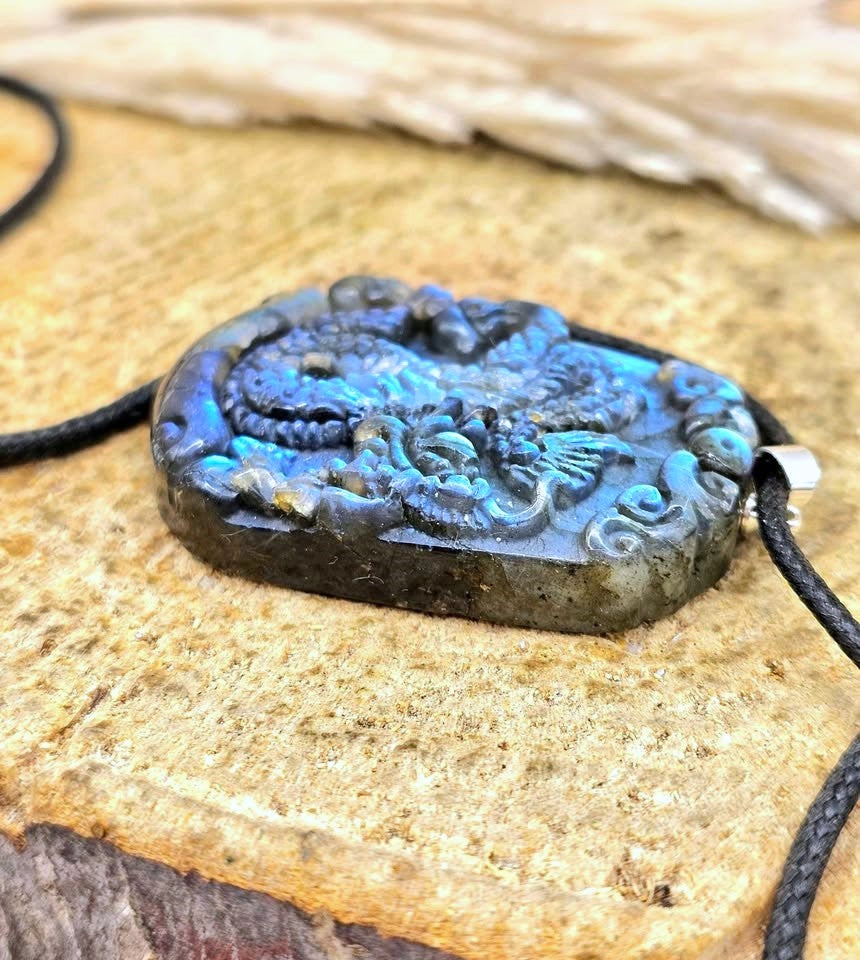 A close-up of a carved oval pendant with a dragon design, featuring vibrant blue and black labradorite stone, hanging from a black cord on a textured tan stone surface.