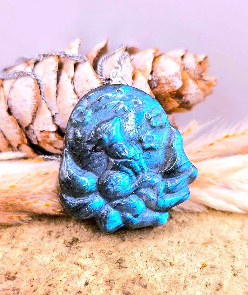 A carved blue stone pendant with a silver bail, resting on a natural pine cone and dried grasses against a soft lavender background.
